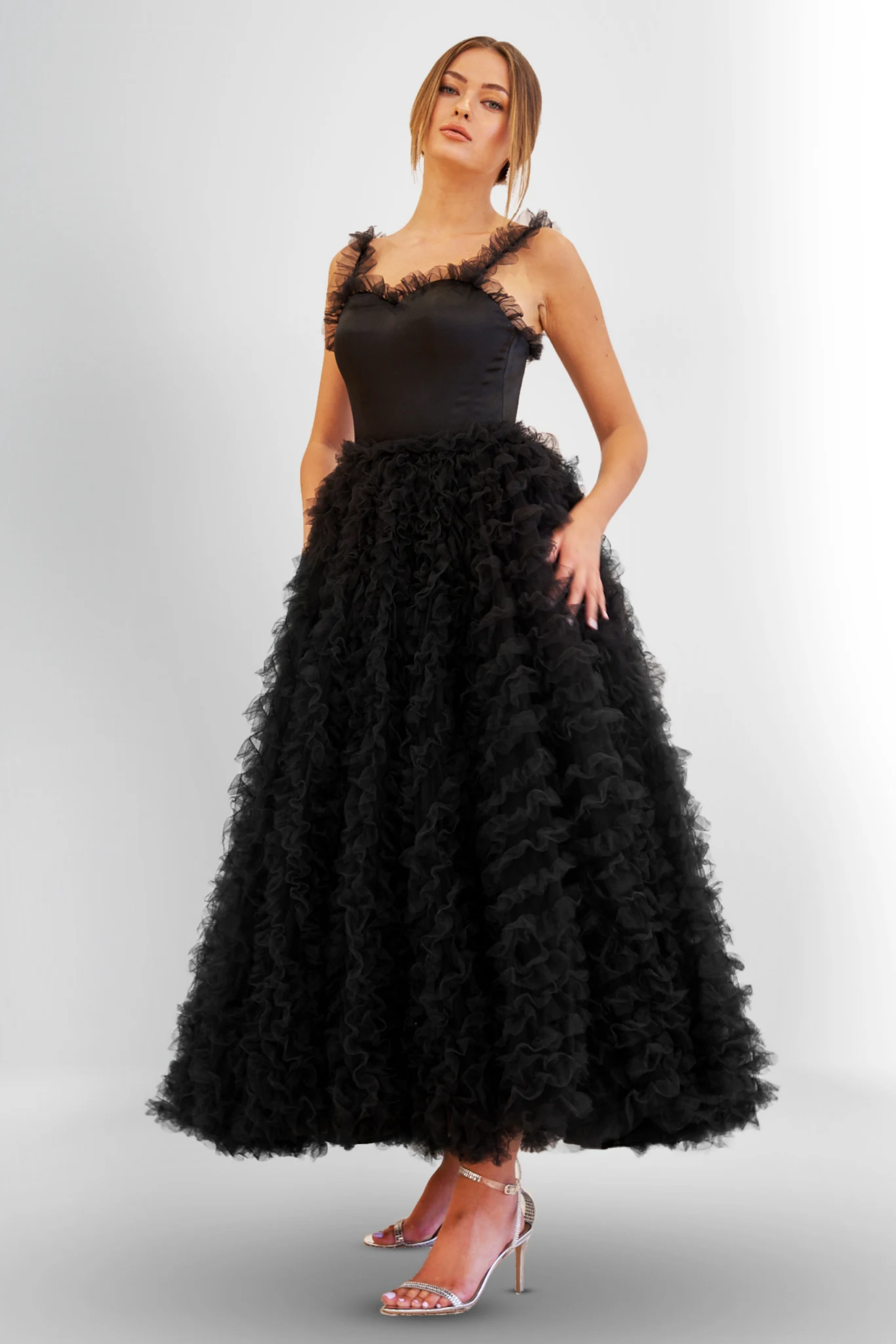 Woman wearing a sleeveless black satin and ruffled tulle midi ball gown with thin straps, paired with silver ankle-strap high heels, posing against a plain light background.