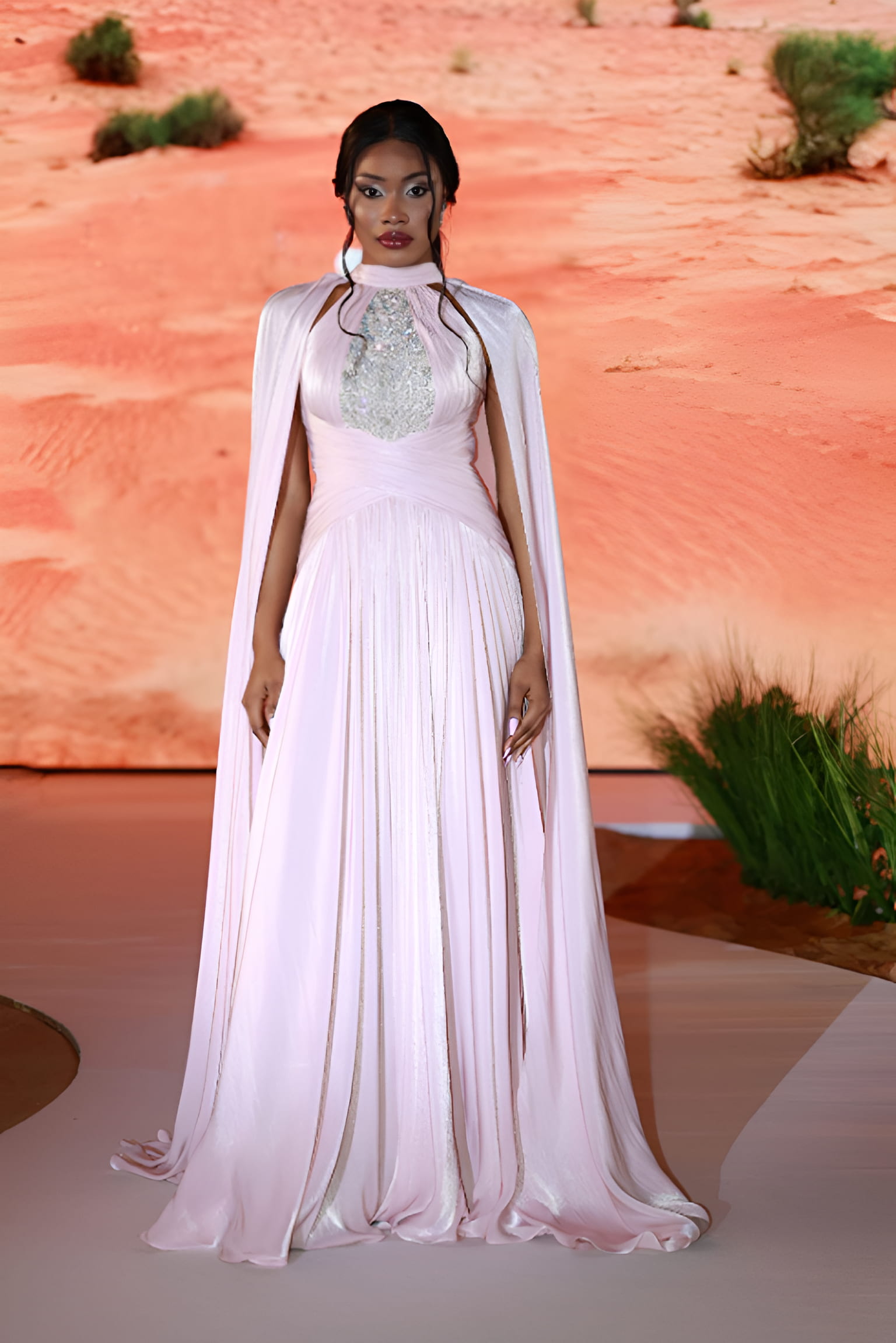 A model wearing a flowing pastel pink gown with a silver embellished bodice and elegant cape, walking on a runway with a desert-inspired backdrop.