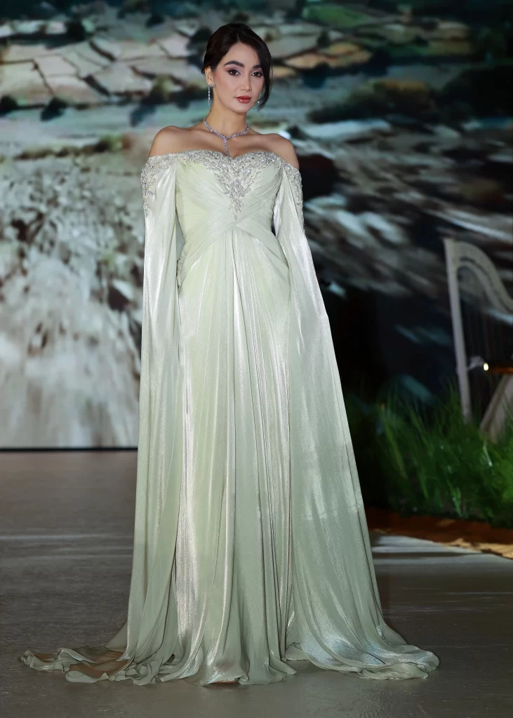 A woman stands elegantly in a shimmering, pale sage off-the-shoulder gown with long, flowing draped sleeves and intricate silver embellishments along the neckline and bodice. The dress cascades to the floor in soft, metallic folds. She wears a delicate necklace and earrings, with her hair styled in an elegant updo. The background shows a blurred, earthy landscape scene.