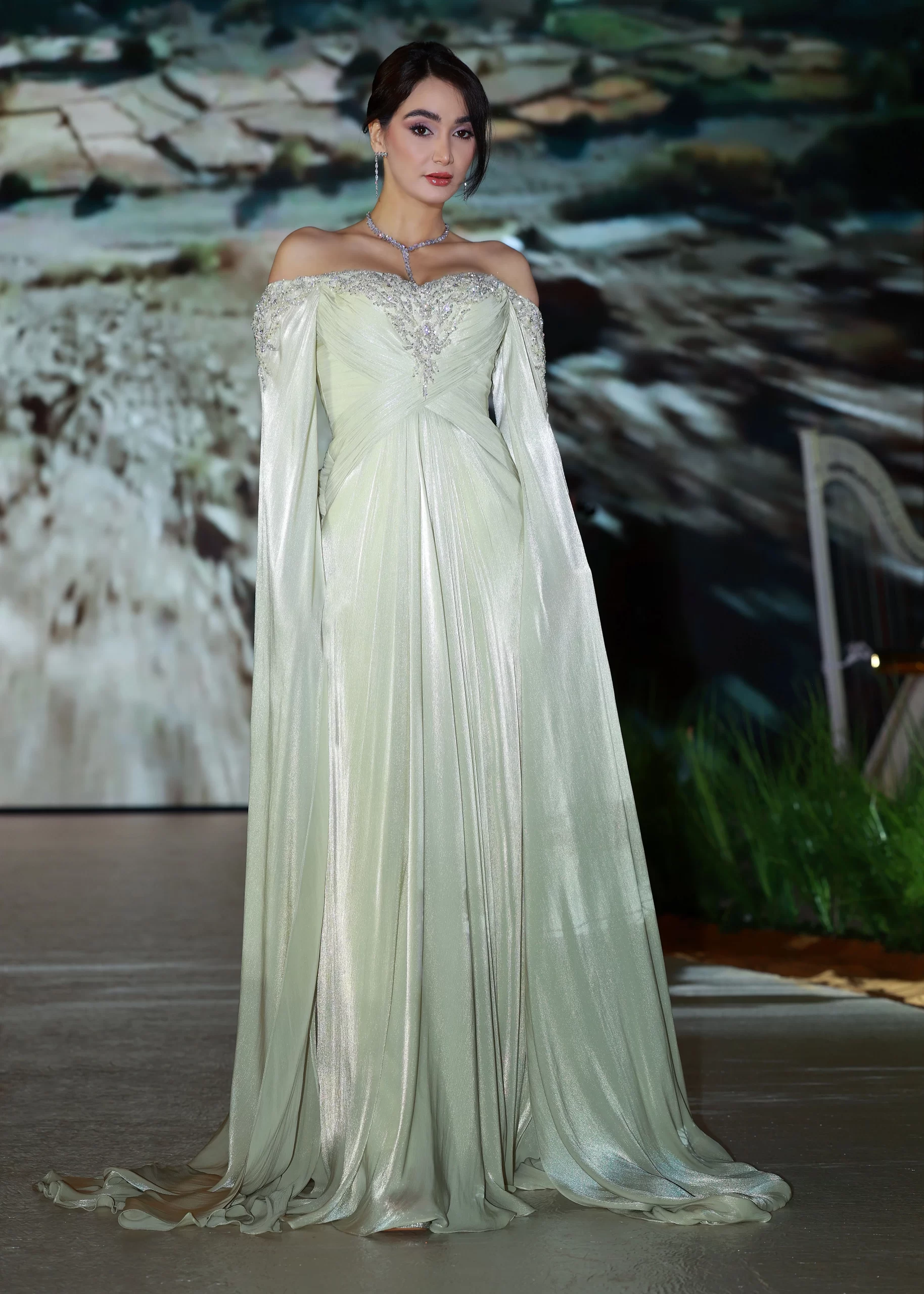 A woman stands elegantly in a shimmering, pale sage off-the-shoulder gown with long, flowing draped sleeves and intricate silver embellishments along the neckline and bodice. The dress cascades to the floor in soft, metallic folds. She wears a delicate necklace and earrings, with her hair styled in an elegant updo. The background shows a blurred, earthy landscape scene.