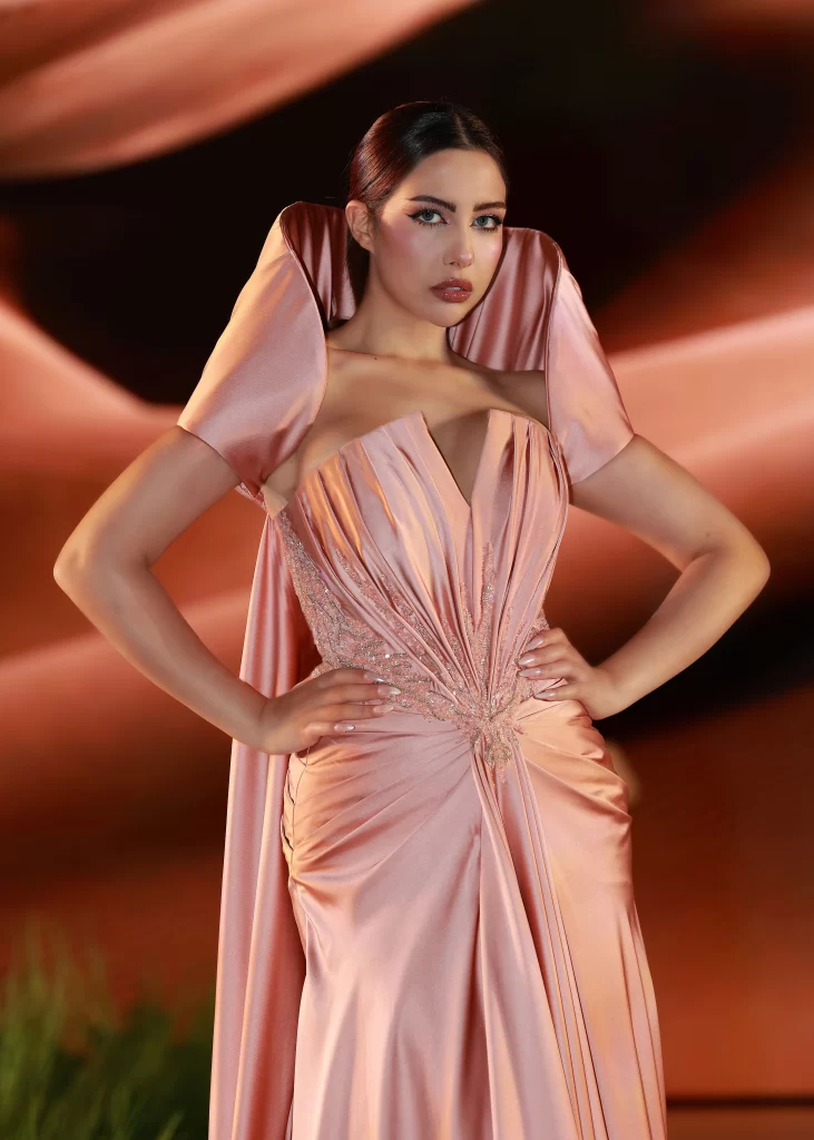 A woman poses confidently in an elegant, shimmering peach-colored gown with dramatic structured shoulders and intricate pleated detailing at the bodice and waist. Her hair is styled sleekly back, and she wears polished makeup with bold eye makeup and glossy lips. The background features warm, flowing, abstract shapes that complement the tones of her dress.