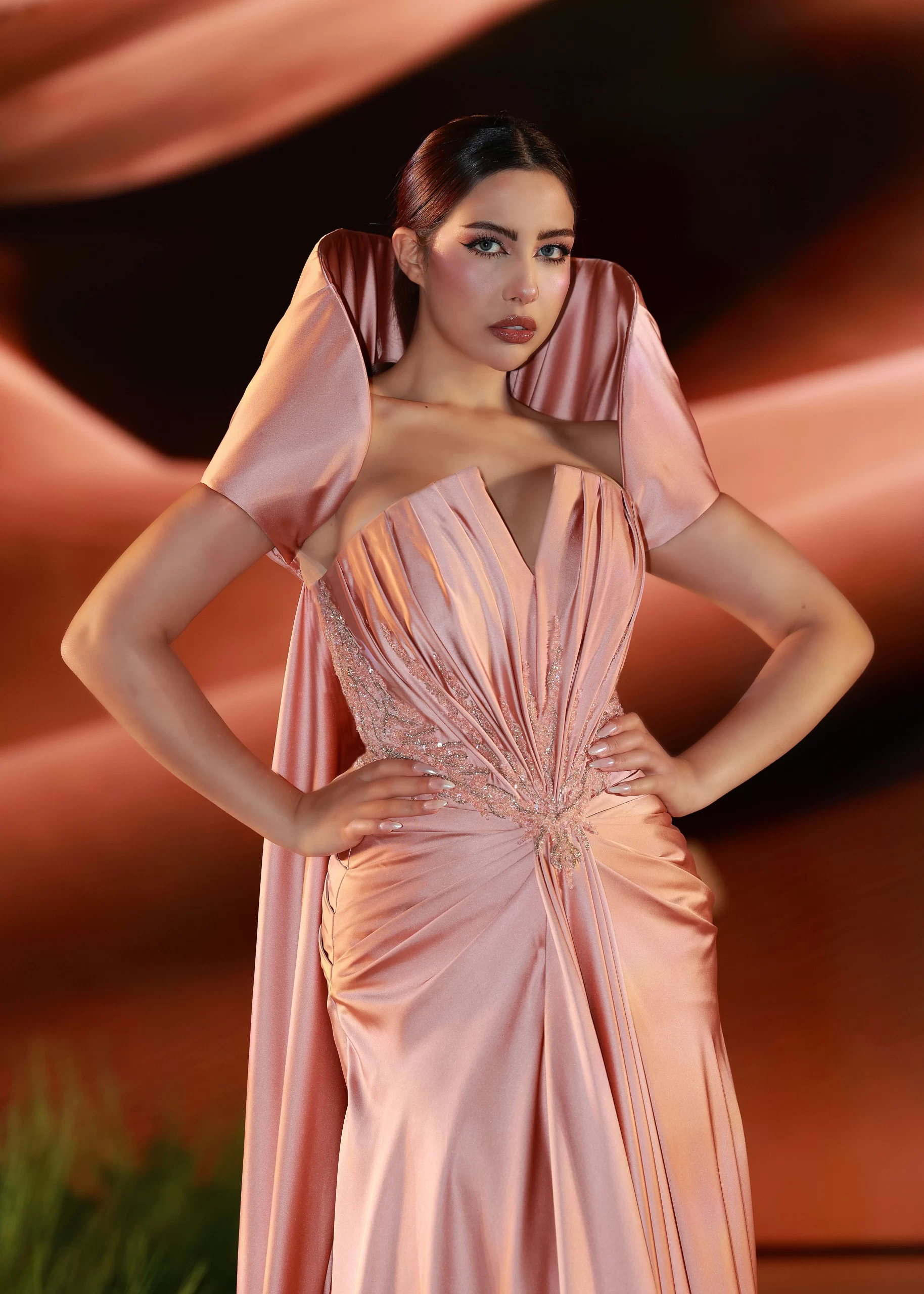 A woman poses confidently in an elegant, shimmering peach-colored gown with dramatic structured shoulders and intricate pleated detailing at the bodice and waist. Her hair is styled sleekly back, and she wears polished makeup with bold eye makeup and glossy lips. The background features warm, flowing, abstract shapes that complement the tones of her dress.
