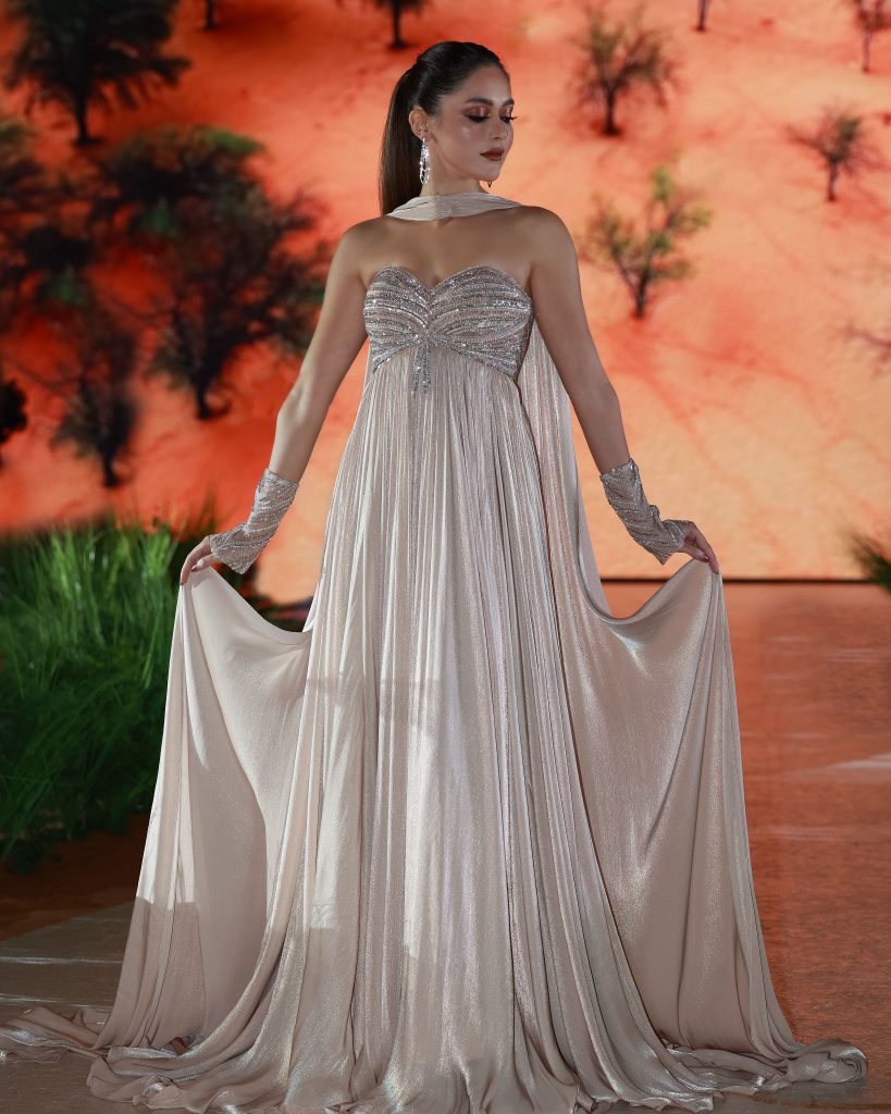 A woman stands elegantly in a shimmering, pale sage off-the-shoulder gown with long, flowing draped sleeves and intricate silver embellishments along the neckline and bodice. The dress cascades to the floor in soft, metallic folds. She wears a delicate necklace and earrings, with her hair styled in an elegant updo. The background shows a blurred, earthy landscape scene.